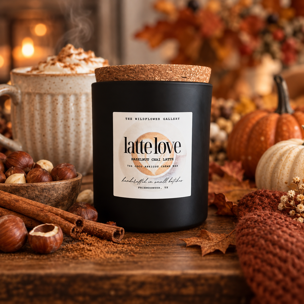 A matte black candle jar with a cork lid and a white label reading 'latte love' and 'The Wildflower Gallery,' styled on a wooden surface with autumn props including pumpkins, cinnamon sticks, hazelnuts, and a mug in the background.